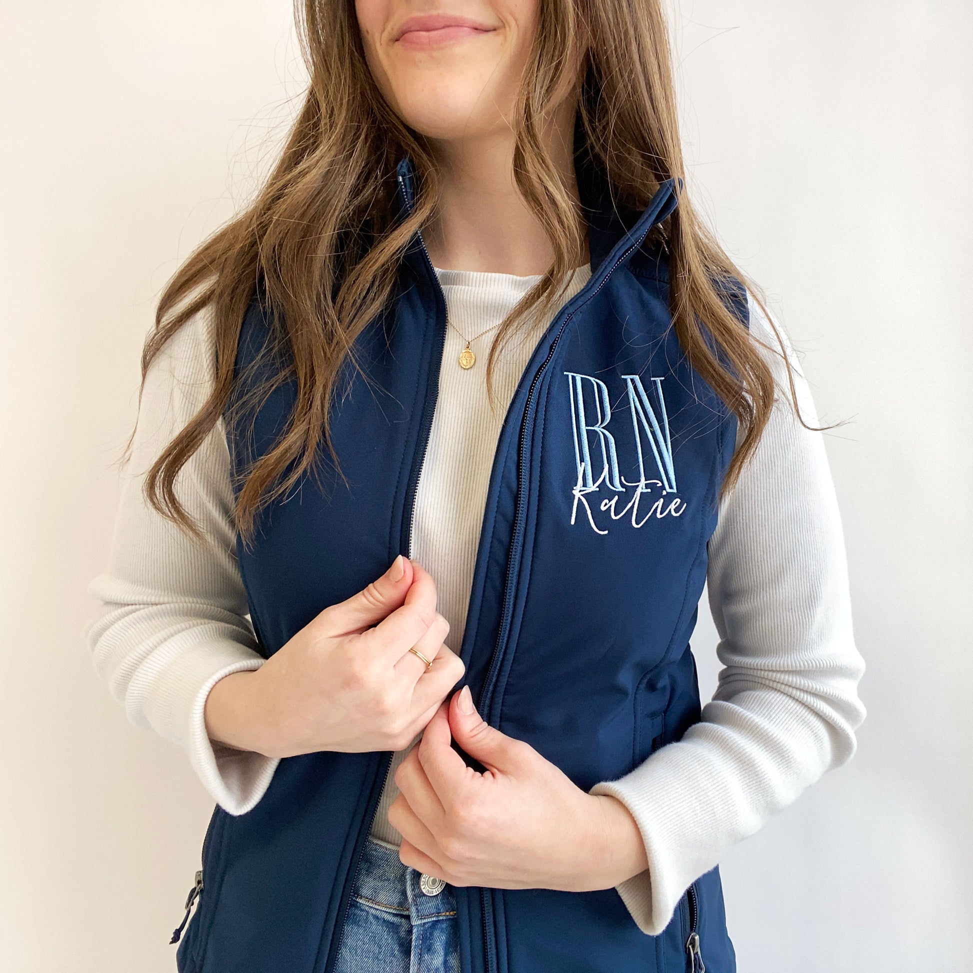 young woman wearing a navy soft shell vest with personalized RN embroidered design on the left chest  in baby blue and white threads