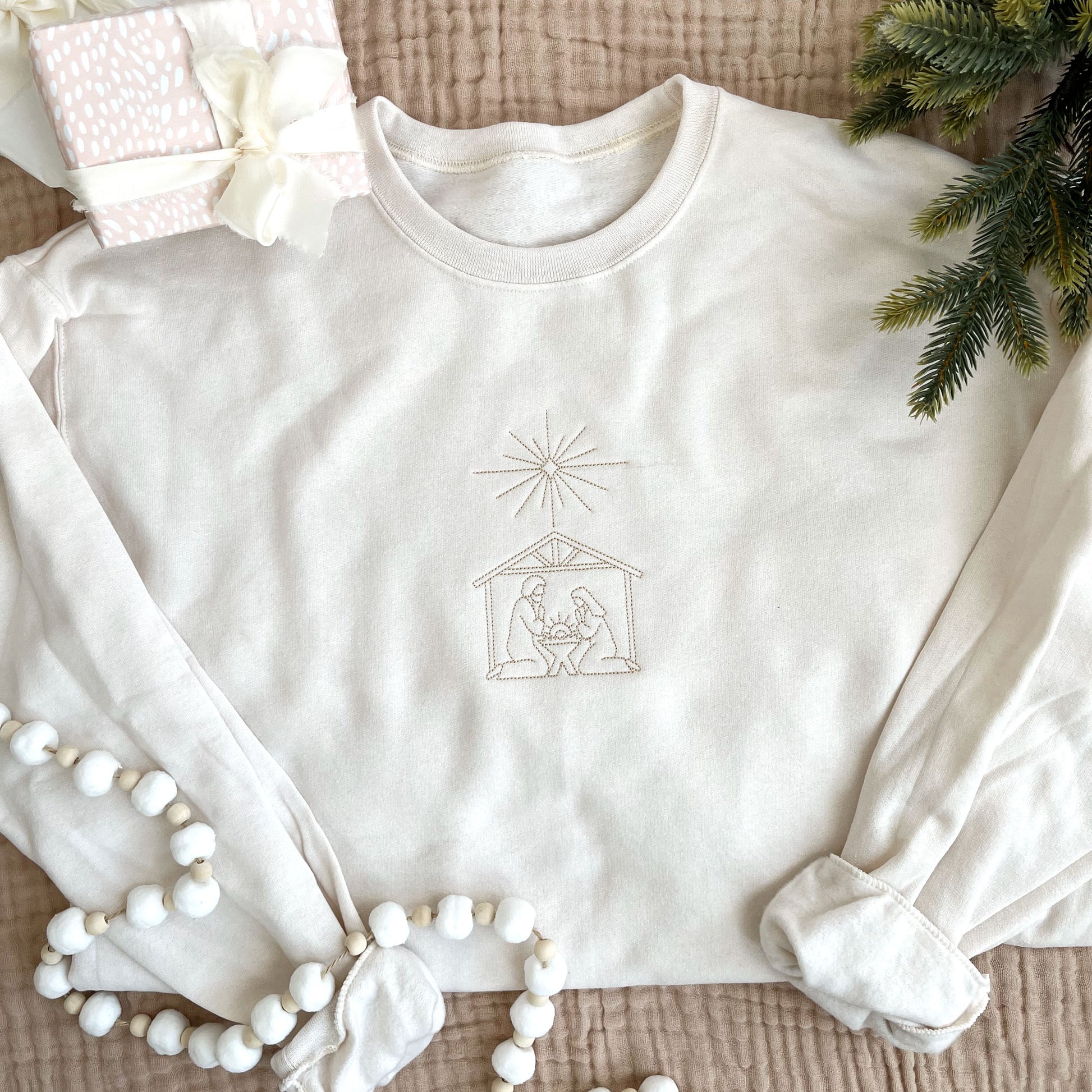 Christmas styled flat lay of a sweet cream crew neck sweatshirt with embroidered stitched nativity scene on the center chest in camel thread.