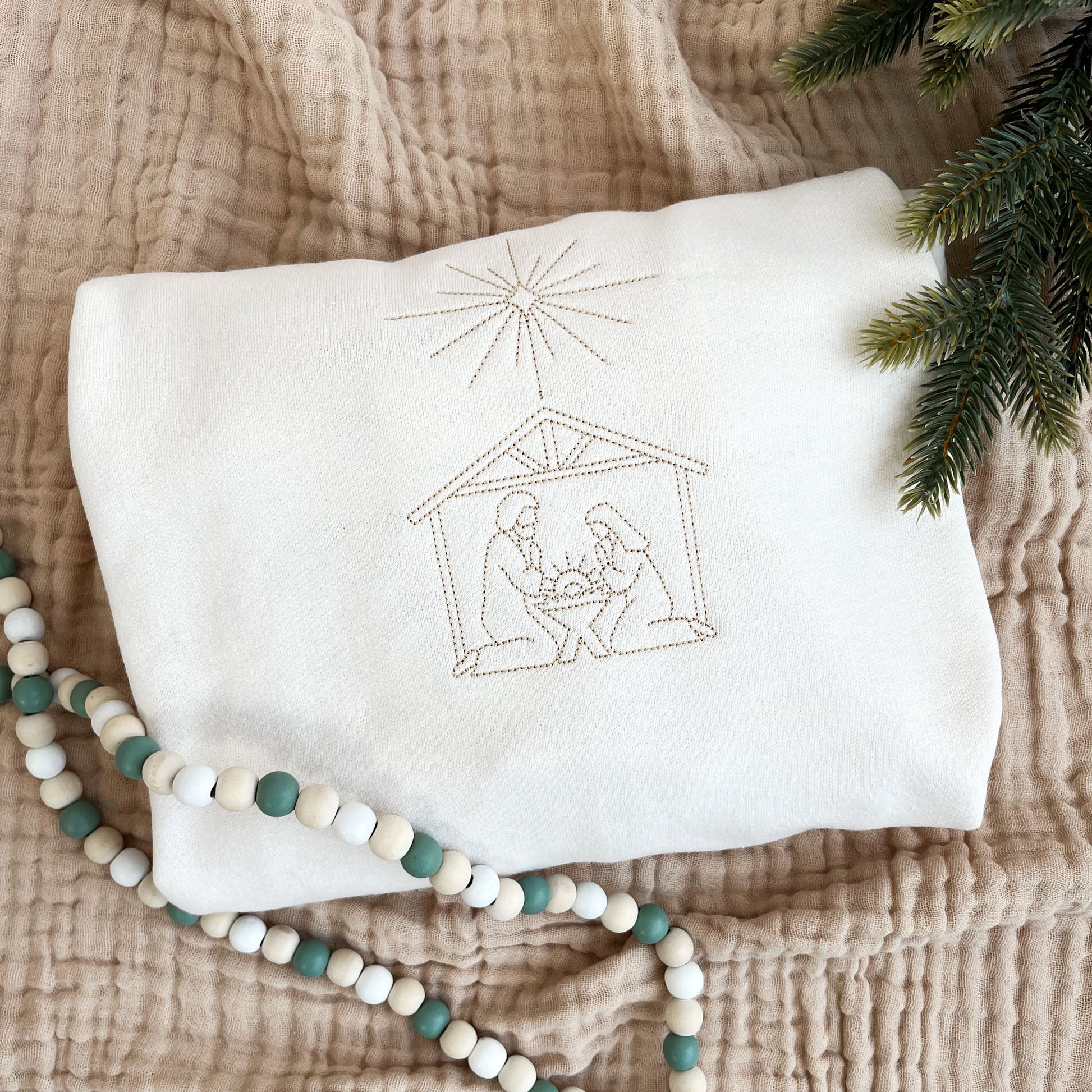 Christmas styled flat lay of a sweet cream crew neck sweatshirt with embroidered stitched nativity scene on the center chest in camel thread.