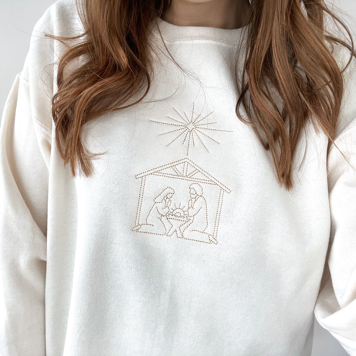 close up of a woman wearing a sweet cream crew neck sweatshirt with embroidered stitched nativity scene on the center chest in camel thread.