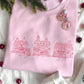 christmas styled flat lay of a light pink crewneck sweatshirt with embroidered stitched christmas village in pink thread