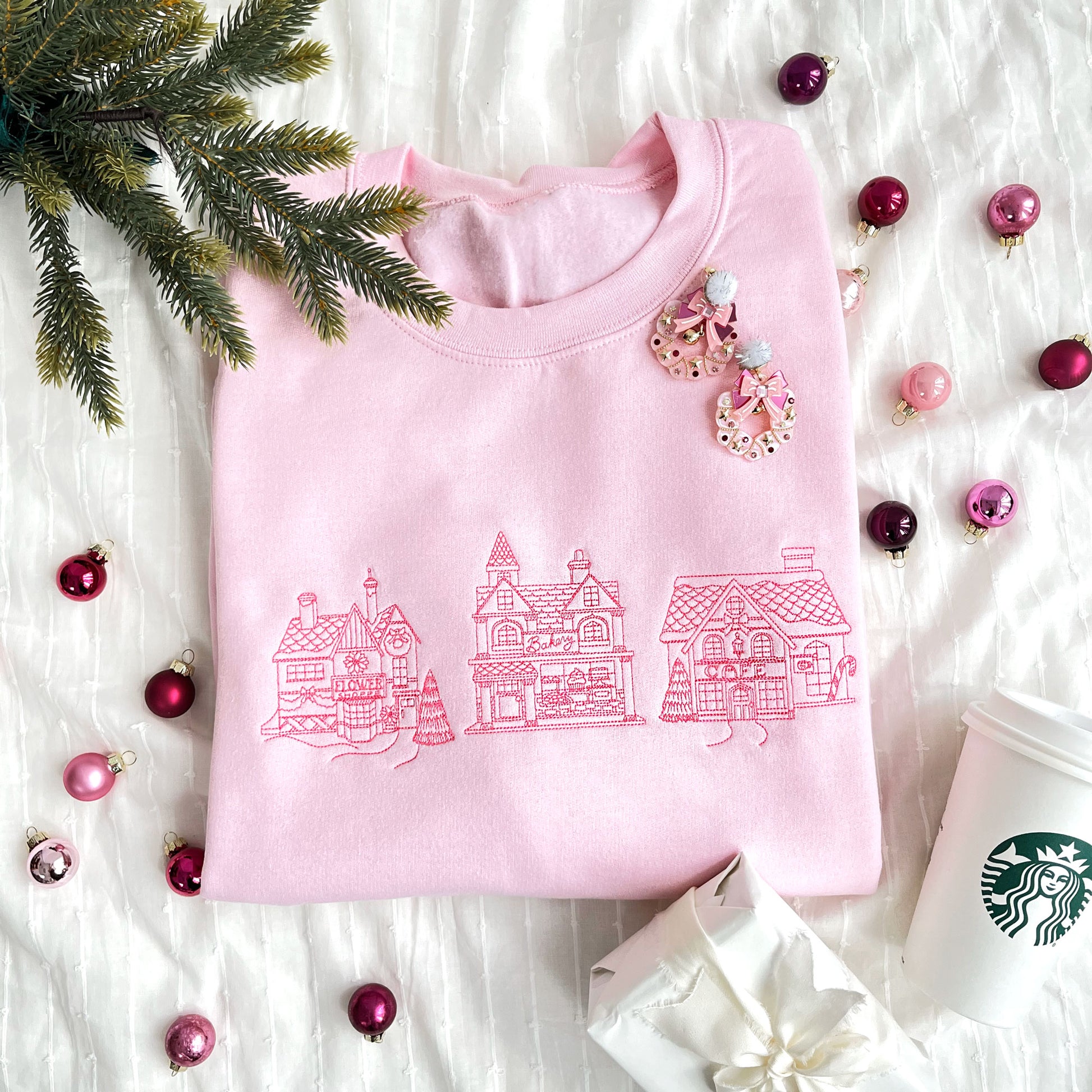 christmas styled flat lay of a light pink crewneck sweatshirt with embroidered stitched christmas village in pink thread