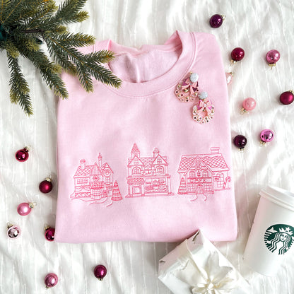 christmas styled flat lay of a light pink crewneck sweatshirt with embroidered stitched christmas village in pink thread