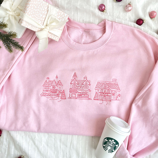 christmas styled flat lay of a light pink crewneck sweatshirt with embroidered stitched christmas village in pink thread