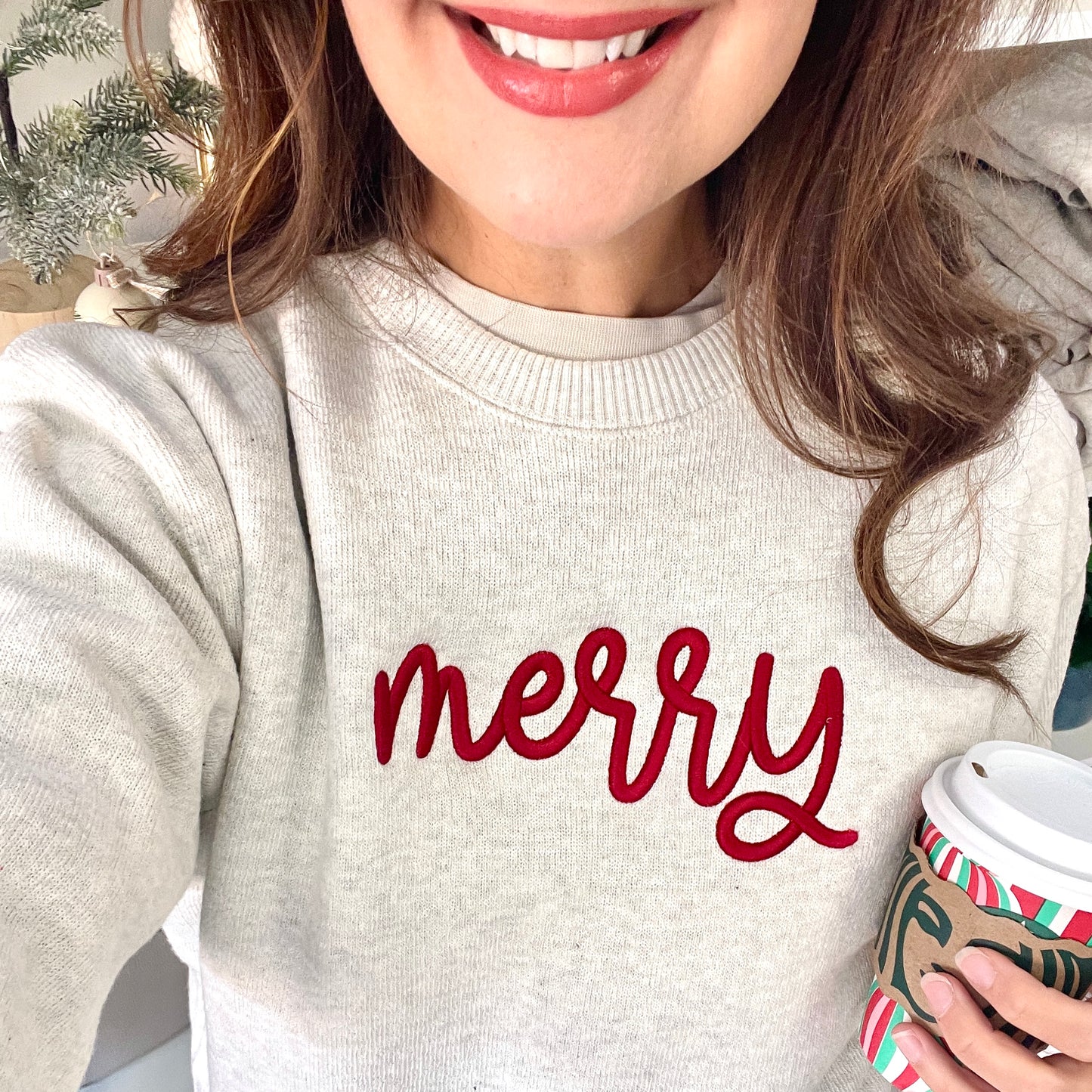 Person wearing a sweater with 'merry' text, holding a coffee cup.