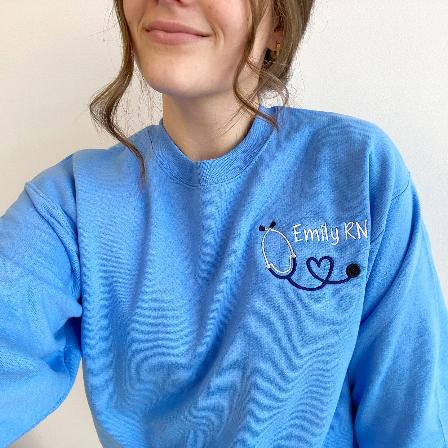 clos up of young woman wearing a Carolina blue crewneck sweatshirt with custom mini heart stethoscope design embroidered on the left chest in navy and white threads
