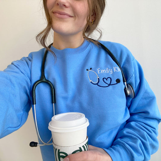 young woman wearing a Carolina blue crewneck sweatshirt with custom mini heart stethoscope design embroidered on the left chest