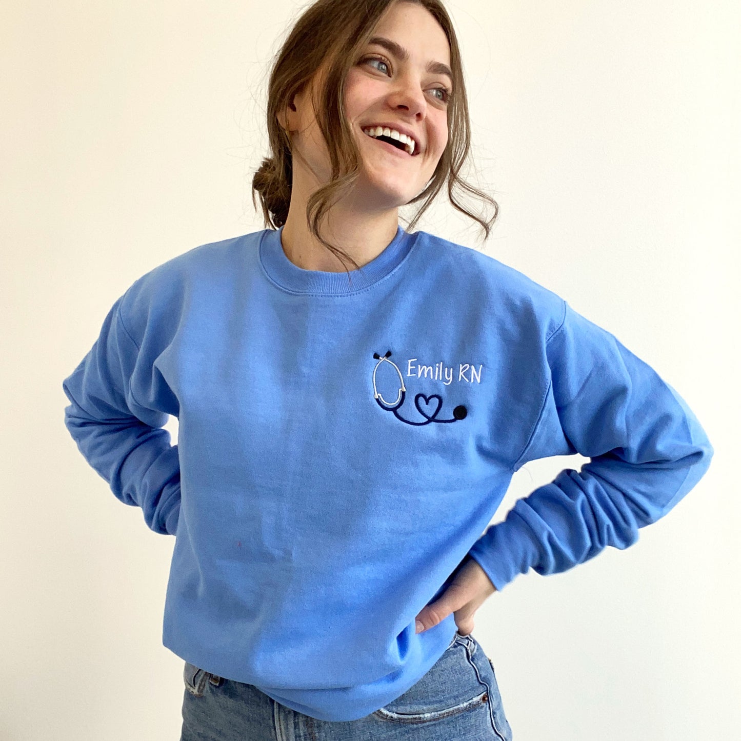 young woman wearing a Carolina blue crewneck sweatshirt with custom mini heart stethoscope design embroidered on the left chest in navy and white threads