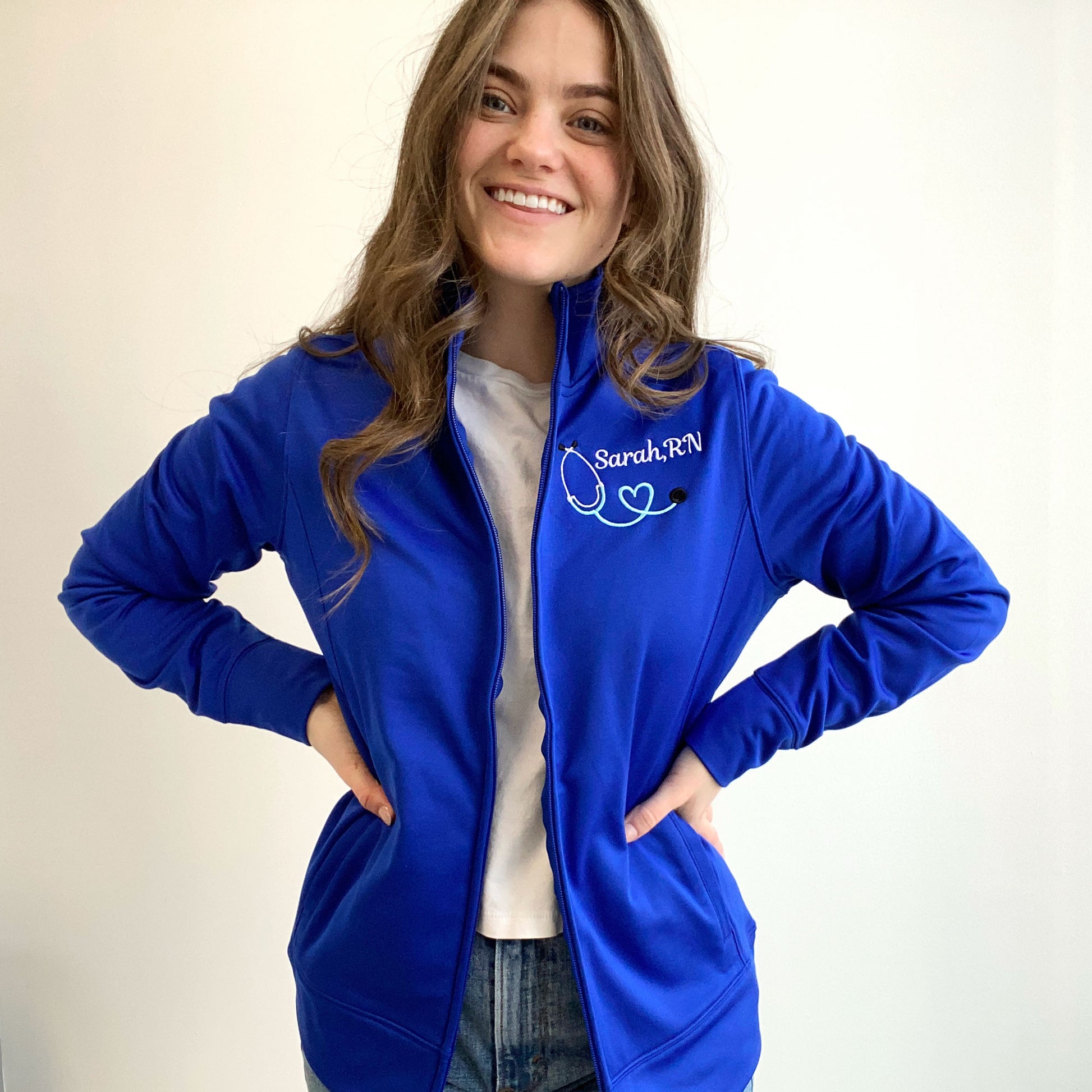 young woman wearing a royal blue full zip polyester nurse jacket with custom embroidered heart stethoscope and name in white and sky thread
