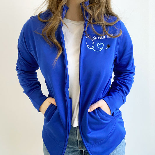 young woman wearing a royal blue full zip polyester nurse jacket with custom embroidered heart stethoscope and name in white and sky thread 