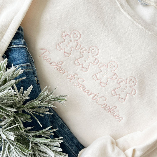 close up of sweet cream crewneck sweatshirt with a custom gingerbread men "teacher of smart cookies" embroidered design on the center chest in powder pink thread