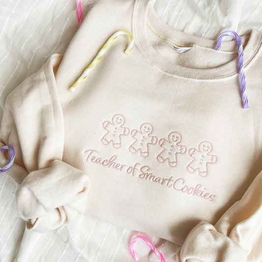 sweet cream crewneck sweatshirt with a custom gingerbread men "teacher of smart cookies" embroidered design on the center chest in powder pink thread