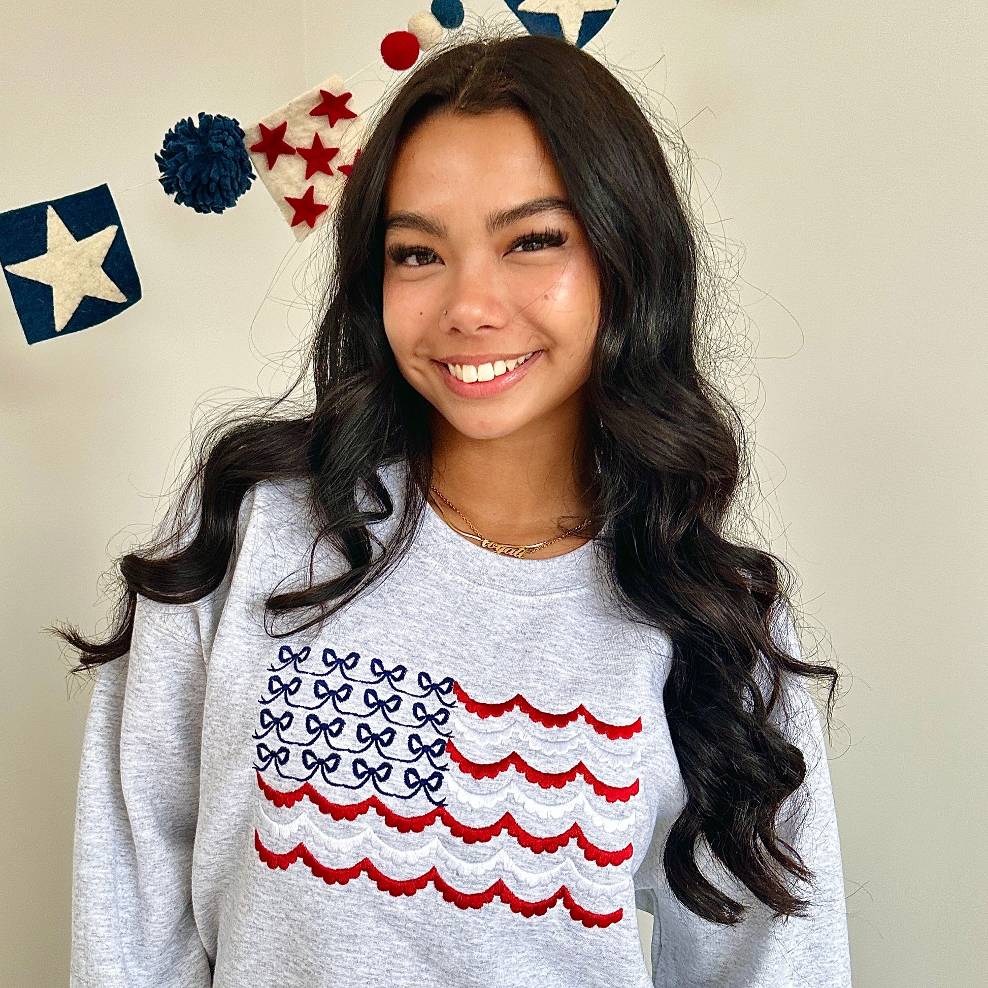 woman with long black hair standing in front of a white wall and patriotic themed wool banner, and she's wearing an oversized ash crewneck sweatshirt with large embroidered American flag design on the front center chest. Flag is designed with navy blue bows instead of stars, and scalloped red and white stripes in place of the traditional stripes on a flag