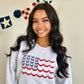 woman with long black hair standing in front of a white wall and patriotic themed wool banner, and she's wearing an oversized ash crewneck sweatshirt with large embroidered American flag design on the front center chest. Flag is designed with navy blue bows instead of stars, and scalloped red and white stripes in place of the traditional stripes on a flag