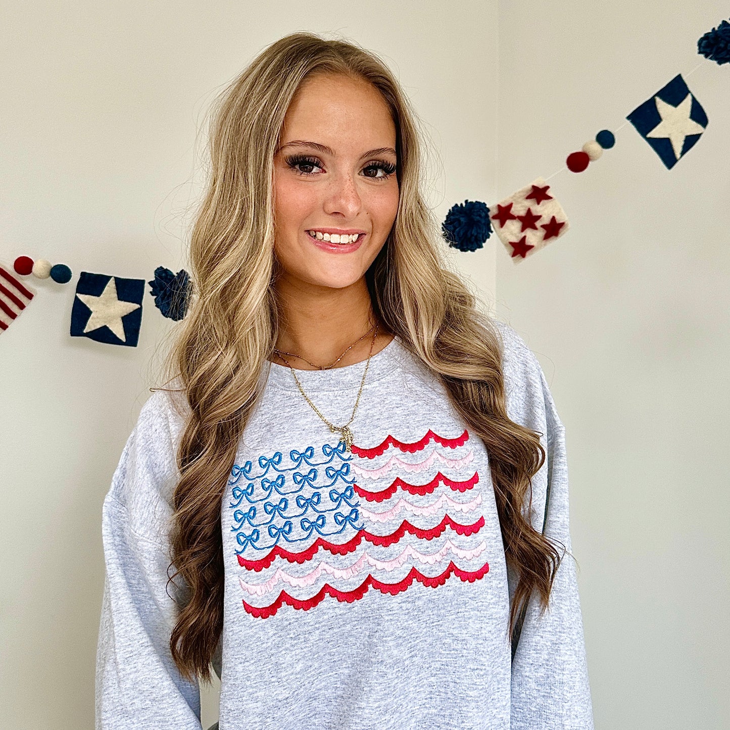 girl wearing an oversized ash crewneck sweatshirt with large embroidered American flag design on the front center chest. Flag is designed with blue bows instead of stars, and scalloped raspberry pink and baby pink stripes in place of the traditional stripes on a flag