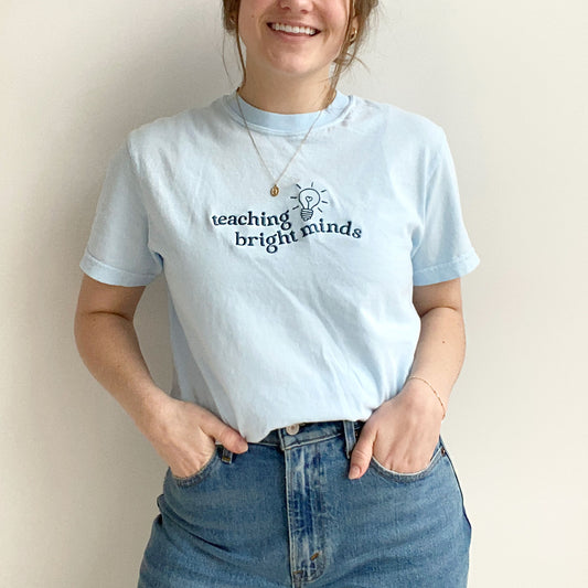 young woman modeling a chambray comfort colors tshirt with teaching bright minds and cute heart lightbulb design embroidered across the chest in french blue threads
