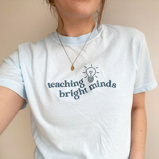 young woman modeling a chambray comfort colors tshirt with teaching bright minds and cute heart lightbulb design embroidered across the chest in french blue threads
