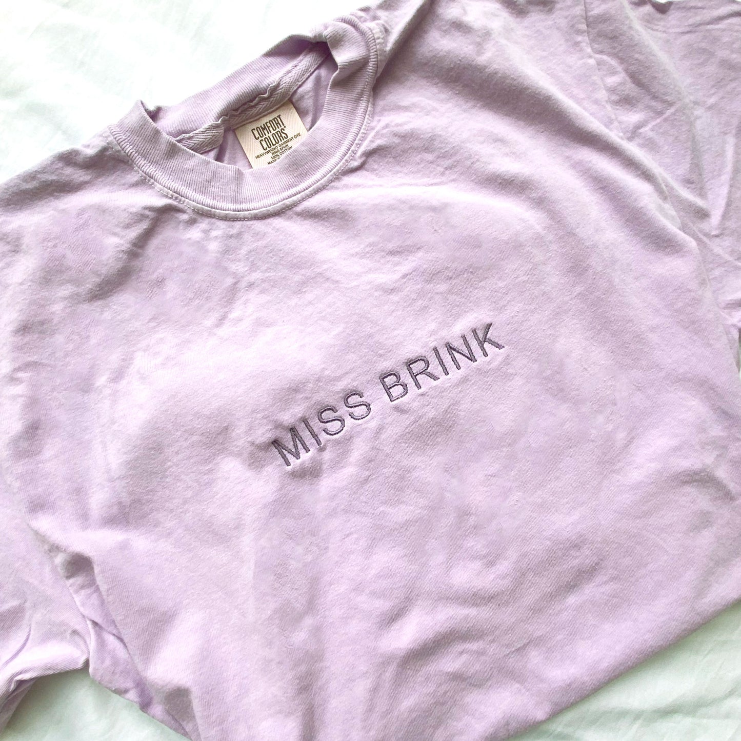 flat lay of an orchid comfort colors t-shirt with minimal custom embroidered name across the chest in smoky orchid thread