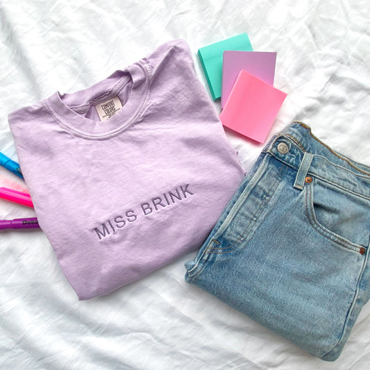 flat lay of an orchid comfort colors t-shirt with minimal custom embroidered name across the chest in smoky orchid thread