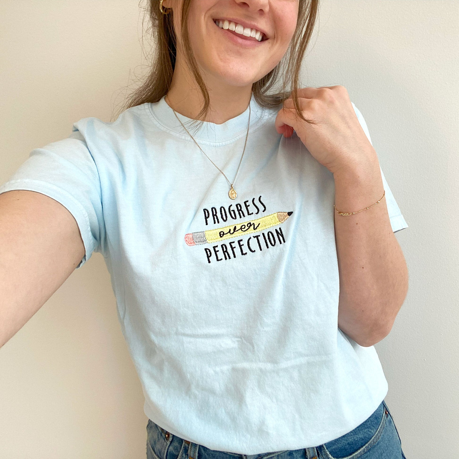 young woman modeling a chambray comfort colors tshirt with embroidered progress over perfection and pencil design 