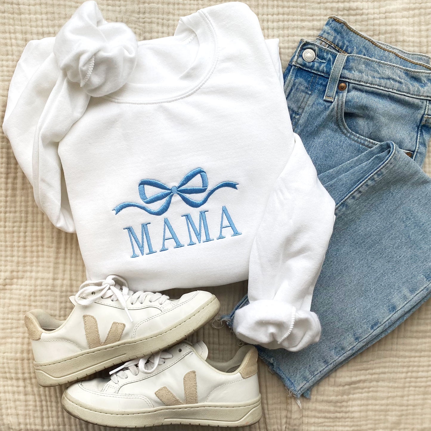 styled flat lay photo of a white crewneck sweatshirt with embroidered bow and all caps mama across the chest in baby blue thread with jeans, jewelry, and sneakers