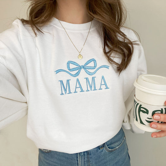 young woman wearing a white crewneck sweatshirt with embroidered large bow and all caps mama design in baby blue thread across the chest holding a Starbucks cup