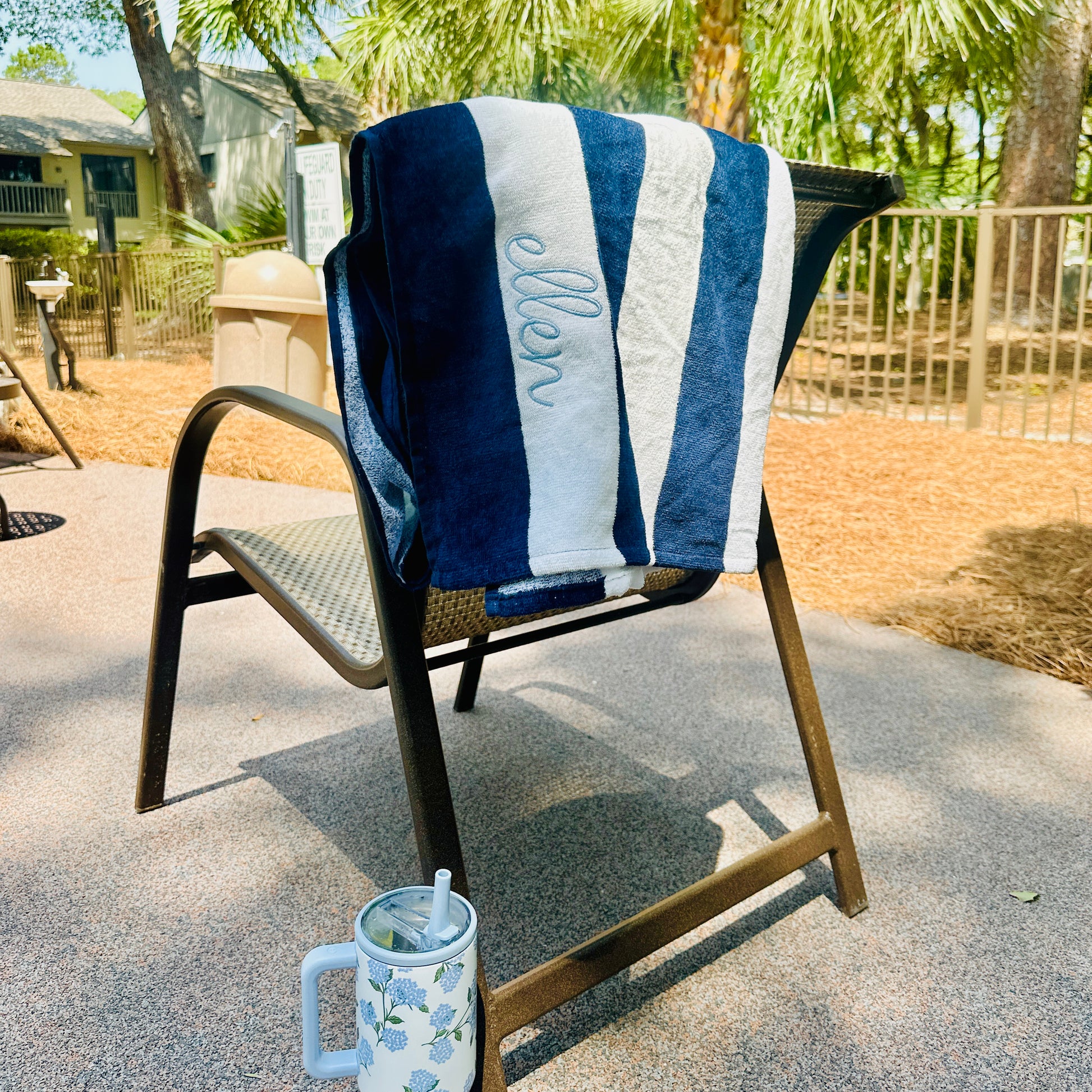navy blue and white striped cabana beach towel with custom name embroidery in a cursive blithe font along the second stripe, horizontally. Embroidery is in powder blue thread. Towel is draped over a chair by the pool. water bottle near by, and palm trees in the background