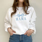 young woman wearing a white crewneck sweatshirt with embroidered large bow and all caps mama design in baby blue thread across the chest
