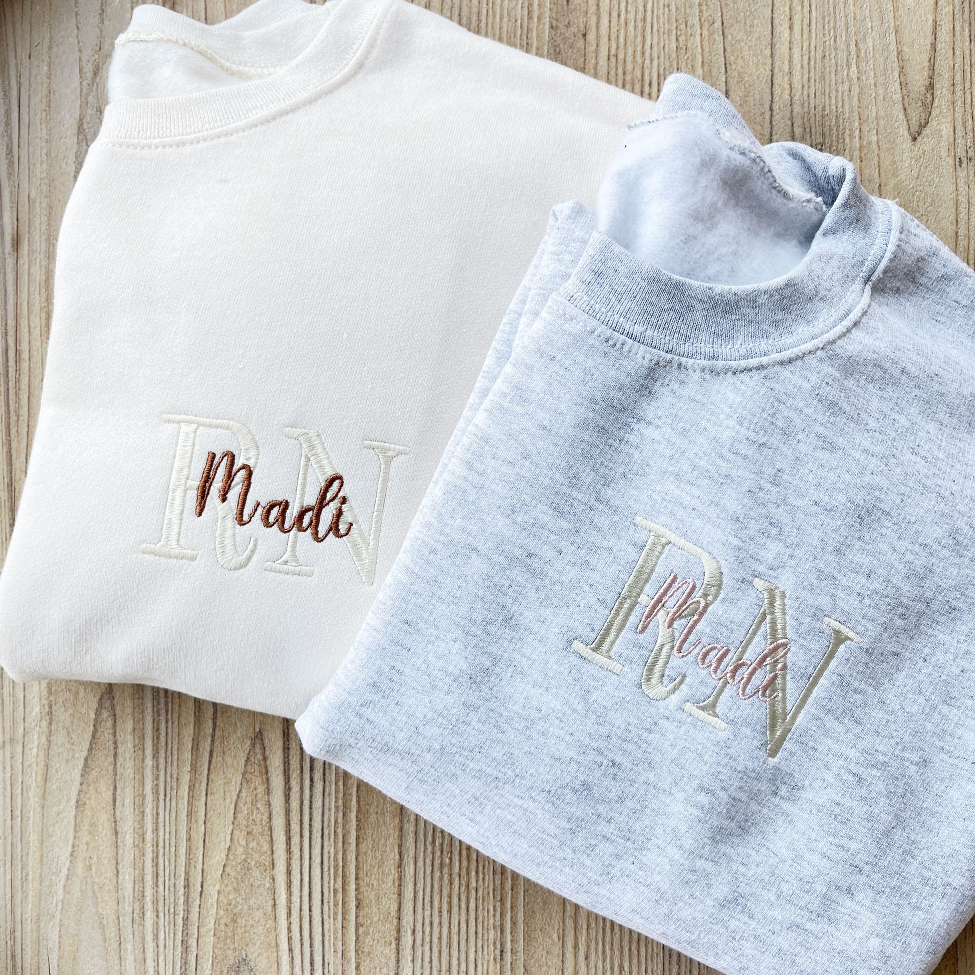 sweet cream crewneck sweatshirt with custom embroidered design RN with name over it in chestnut and natural thread and an ash crewneck sweatshirt with custom embroidered design RN with name over it in natural and powder pink thread