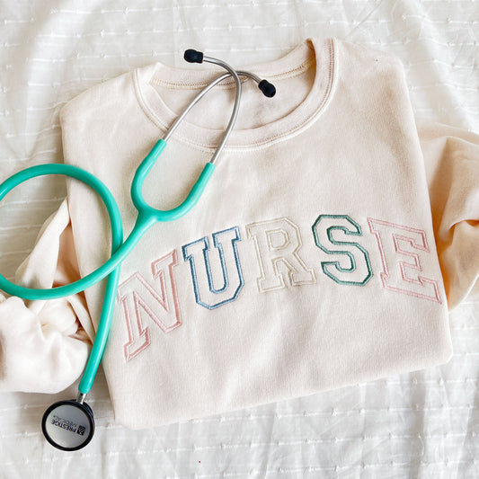 sweet cream crewneck sweatshirt with custom NURSE athletic outlined font embroidered in pastel pink, purple, natural, and eucalyptus thread