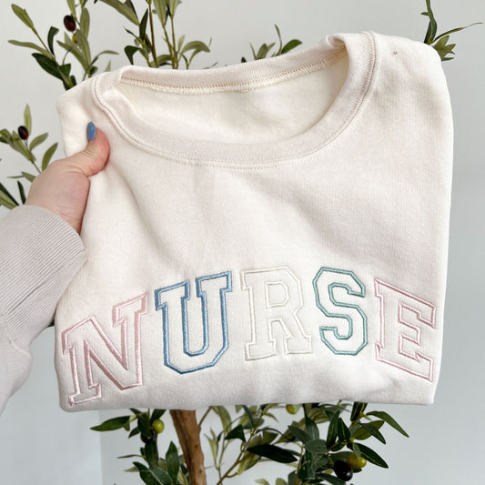 sweet cream crewneck sweatshirt with custom NURSE athletic outlined font embroidered in pastel pink, purple, natural, and eucalyptus thread