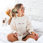 young woman sitting on a bed holding coffee wearing a sweet cream crewneck sweatshirt with embroidered coffee club across the chest in chestnut  thread and schoolbook lower case font