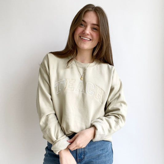 young woman wearing a sand crewneck sweatshirt with embroidered neutral color block teach design across the chest