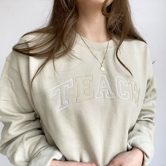 young woman wearing a sand crewneck sweatshirt with embroidered neutral color block teach design across the chest