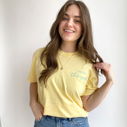 young woman wearing a butter comfort colors short sleeve tshirt with embroidered speech therapist design on the left chest in sky blue thread