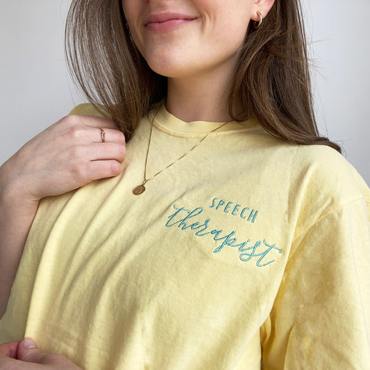 close up of a young woman wearing a butter comfort colors short sleeve tshirt with embroidered speech therapist design on the left chest in sky blue thread