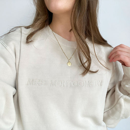 young woman modeling an ivory crewneck sweatshirt with mininmal embroidered name in all caps across the chest in natural thread