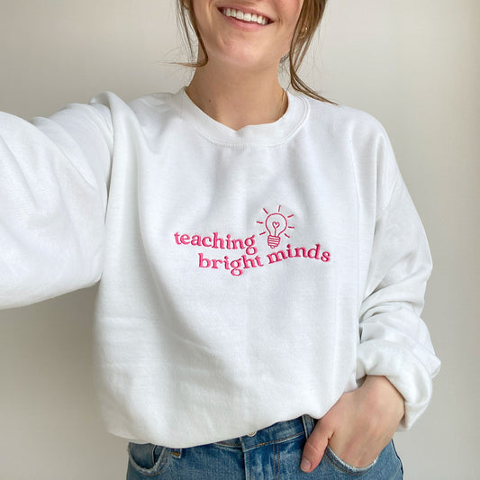 young woman wearing a white teacher pullover sweatshirt with a teaching bright minds lightbulb embroidered design on the chest