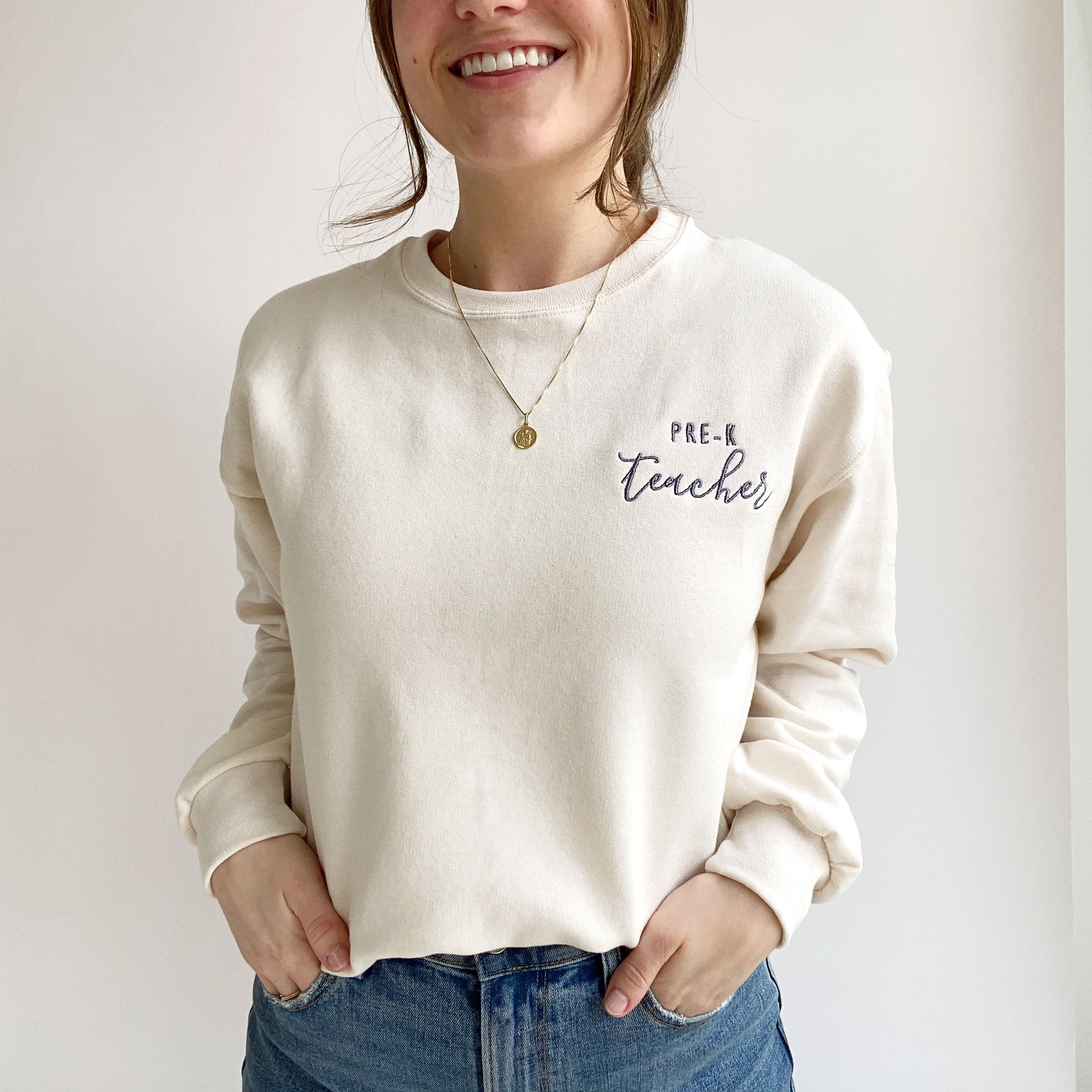 young woman wearing a sweet cream crewneck sweatshirt with custom teacher grade design embroidered in smoky orchid thread
