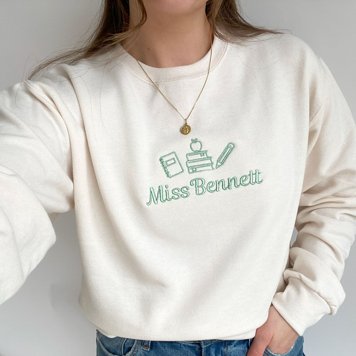 close up of a young woman wearing a sweet cream crewneck sweatshirt with small embroidered teacher icons and custom name in silver sage thread across the chest