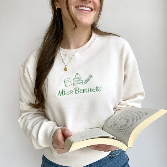 young woman wearing a sweet cream crewneck sweatshirt with small embroidered teacher icons and custom name in silver sage thread across the chest