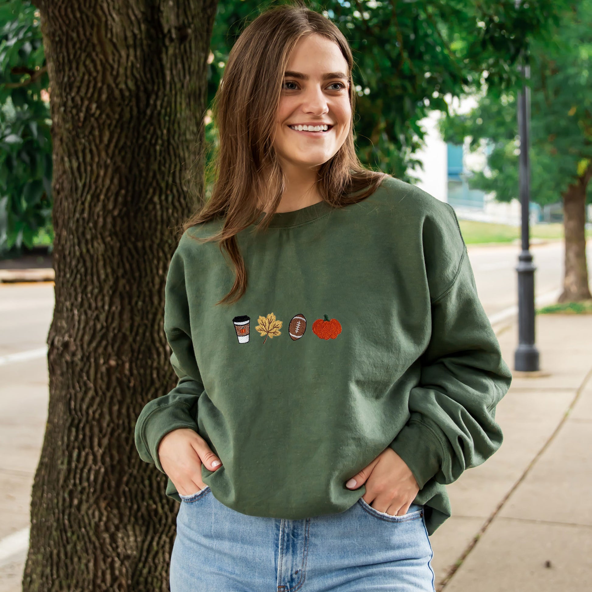 close up of a woman wearing a military green crewneck sweatshirt with embroidered coffee, leaf,  ootball, and pumpkin design across the chest