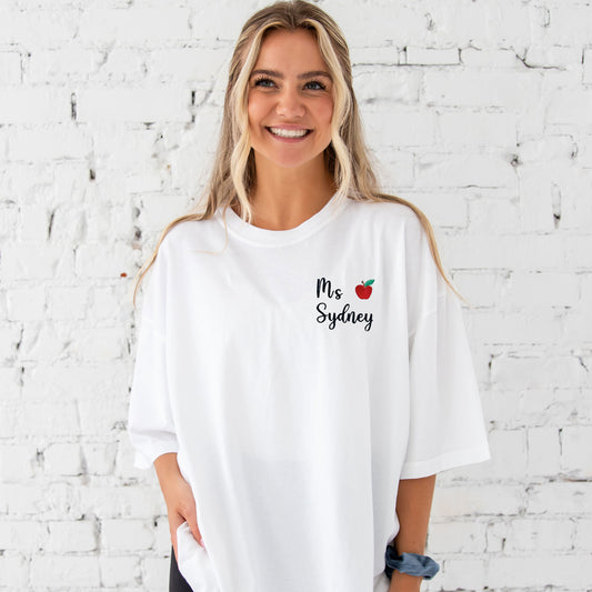 young woman wearing a white comfort colors t-shirt with embroidered name and mini apple embroidered on the left chest