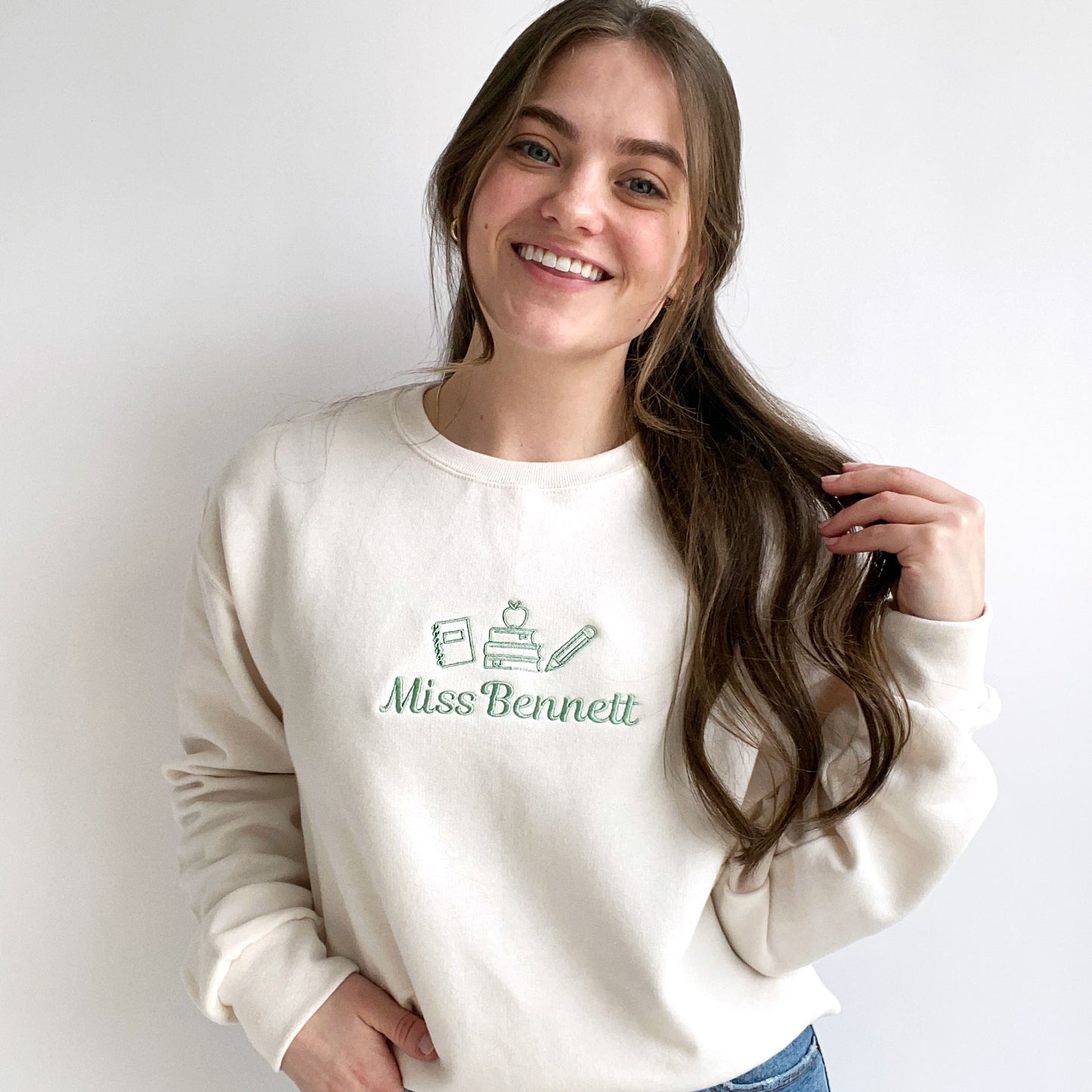 young woman wearing a sweet cream crewneck sweatshirt with small embroidered teacher icons and custom name in silver sage thread across the chest