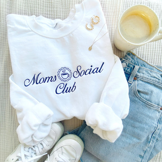 white crewneck sweatshirt styled with jeans and sneakers and a coffee cup. On the sweatshirt is embroidered Moms Social club design with little coffee cup in navy threads