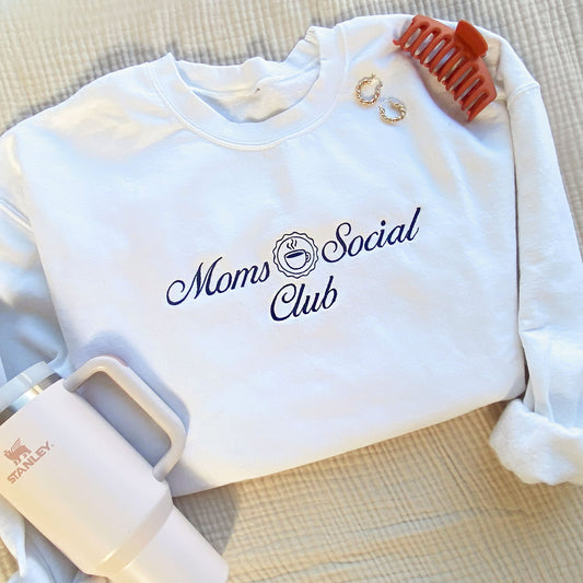 flat lay of a white crewneck sweatshirt with embroidered Moms Social club design in navy thread and styled with a Stanley cup and a claw clip with earrings