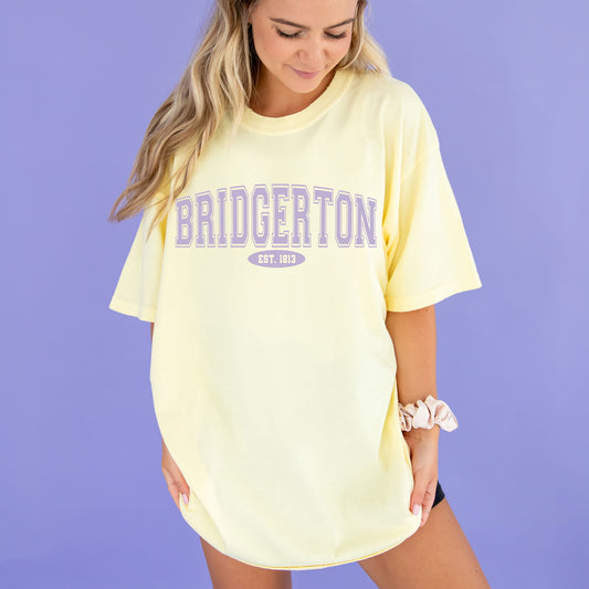 woman wearing an oversized yellow tee with a varsity inspired BRIDGERTON print in purple.