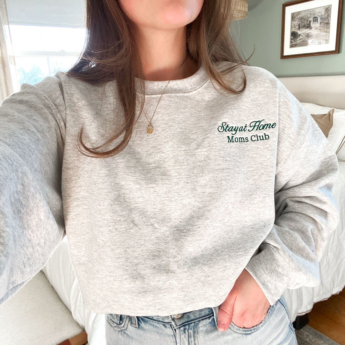  young woman wearing an ash crewneck sweatshirt with embroidered stay at home moms cub design on the left chest in hunter green thread