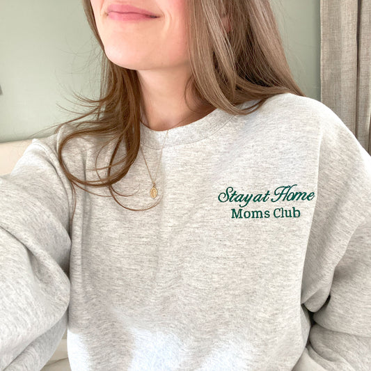 close up of a young woman wearing an  ash crewneck sweatshirt with embroidered stay at home moms cub design on the left chest