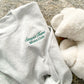 close up image of a flat lay of an ash crewneck sweatshirt with embroidered stay at home moms cub design on the left chest in hunter green thread styled with fuzzy slippers